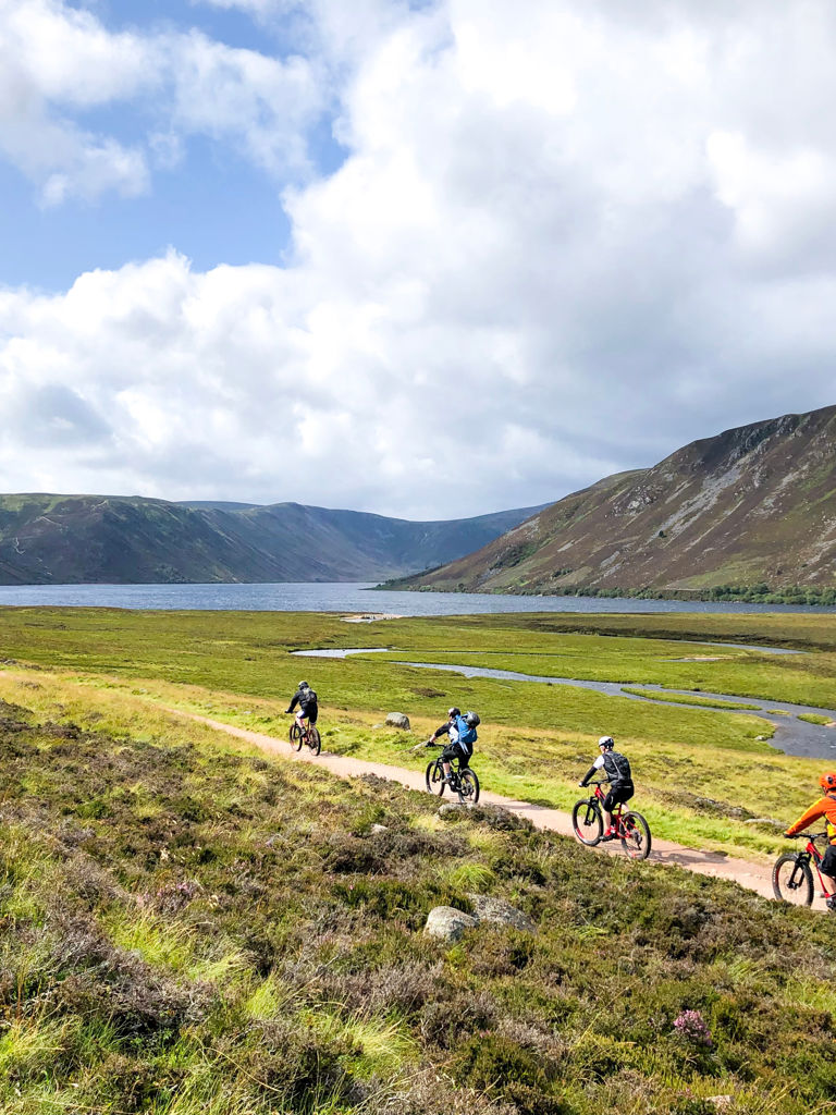 Group of people riding bicycles near a lake, lochnagar, during daytime, mountains appear in the background Group of people riding bicycles near a lake, lochnagar, during daytime, mountains appear in the background