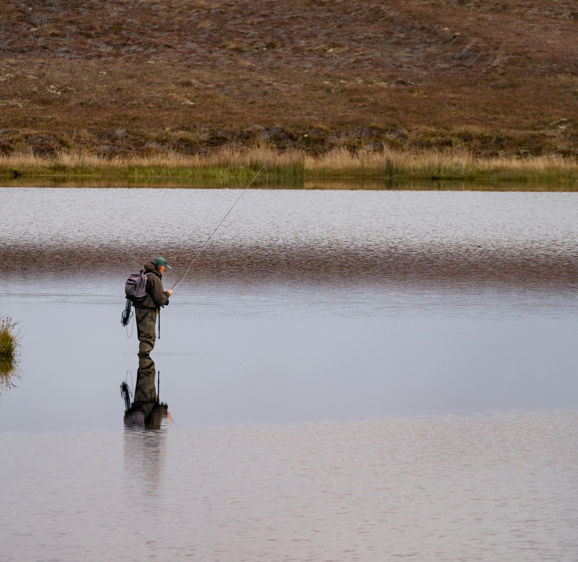 Fisherman alone stand in river water. Hobby sport activity. Fisherman fishing equipment. Reflection in Loch Long. Loch Long Fisherman alone stand in river water. Hobby sport activity. Fisherman fishing equipment. Reflection in Loch Long. Loch Long